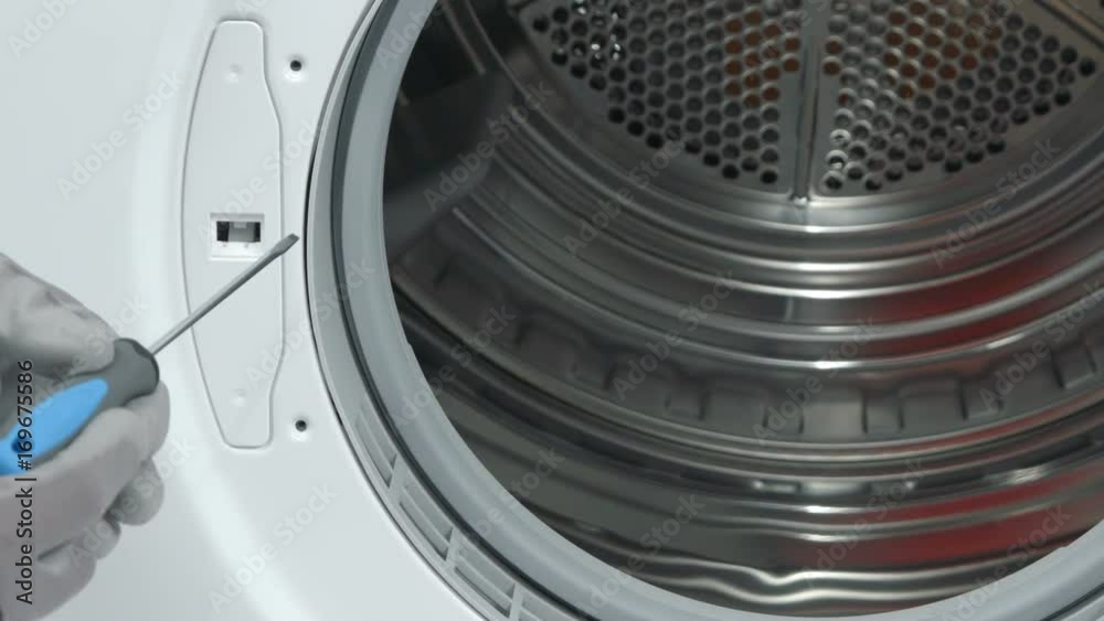 A repairman is taking down a cover next to a drum in a washing machine. He is fixing the device with a screwdriver. 