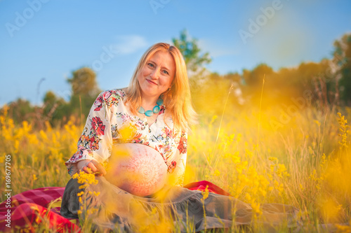 9 months pregnant woman with mehandi pattern ornament belly sitting in yellow grass and smiling on sunset lights. Waiting for baby. Pregnancy concept