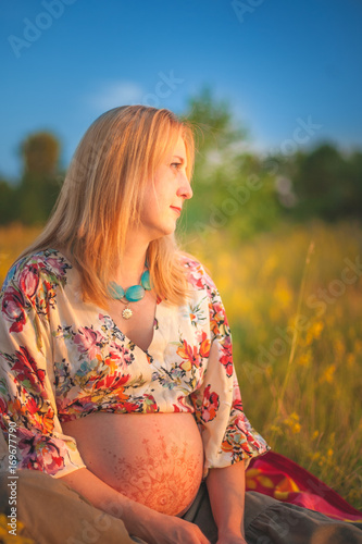 9 months pregnant woman with mehandi pattern ornament belly sitting in yellow grass and smiling on sunset lights. Waiting for baby. Pregnancy concept