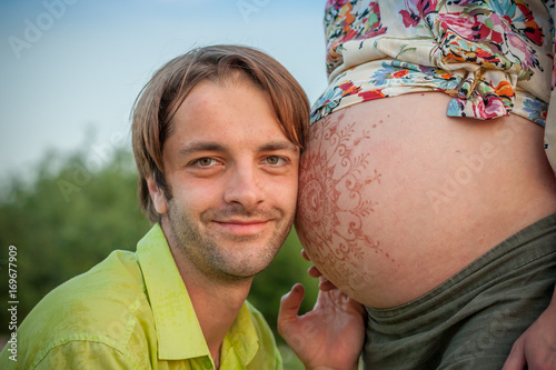 Happy husband littening his wifes pregnant mehandi pattern ornament belly on sunset nature landscape