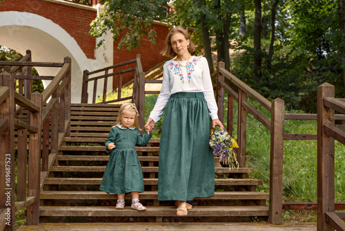  Family mom with daughter in vintage retro style linen dresses  with bouquet of flowers walking dawn wooden stairs in a park garden