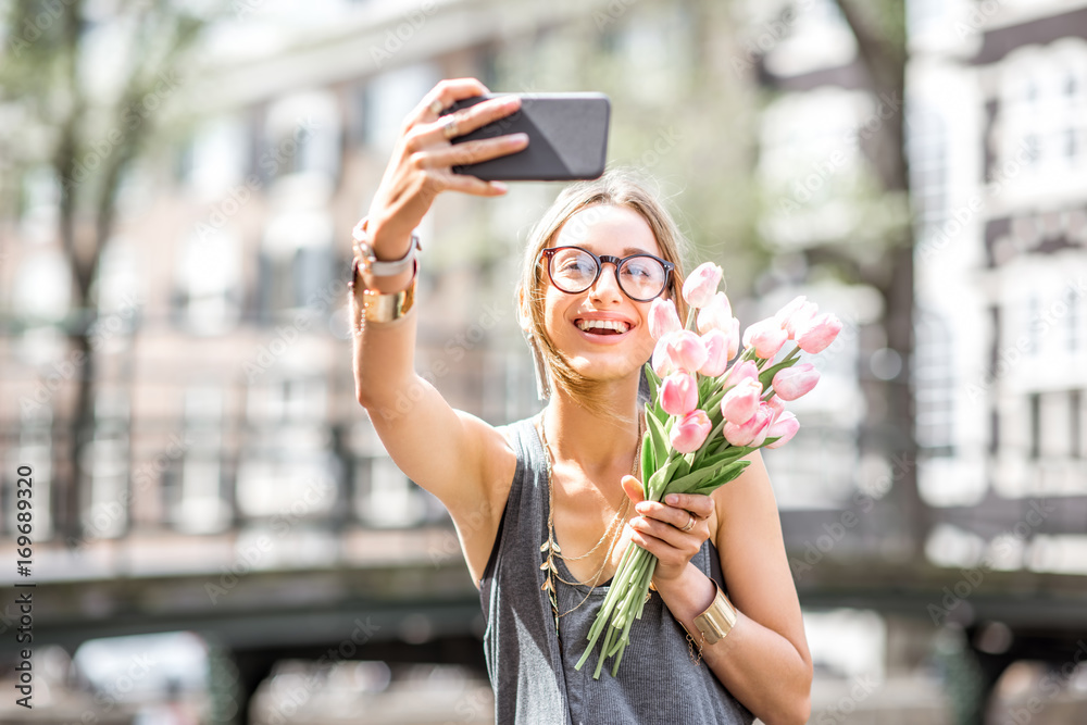 Fototapeta premium Young woman making selfie photo with bouquet of pink tulips sitting on the fence near the water channel in Amsterdam city