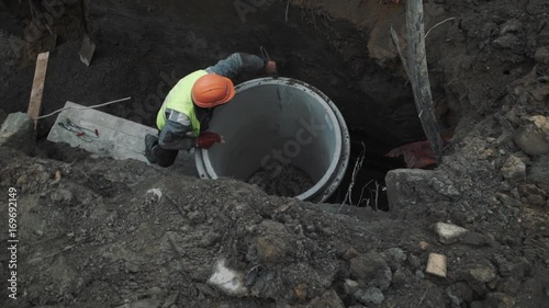 Worker in orange hard hat unscrew metal noose of concrete chamber manhole ring in a ditch at sewer building site