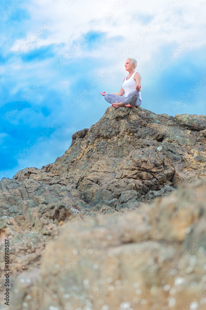 Beautiful mature aged woman sits on top of a cliff doing yoga