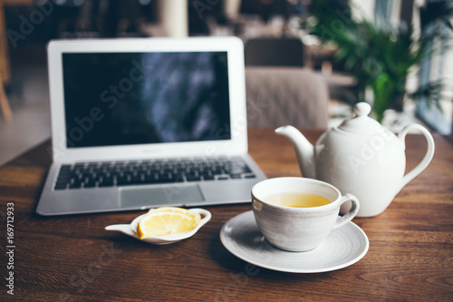 Close-up shoot of cup of hot tea with lemon and infusion teapot on wooden table with laptop in modern cafe 