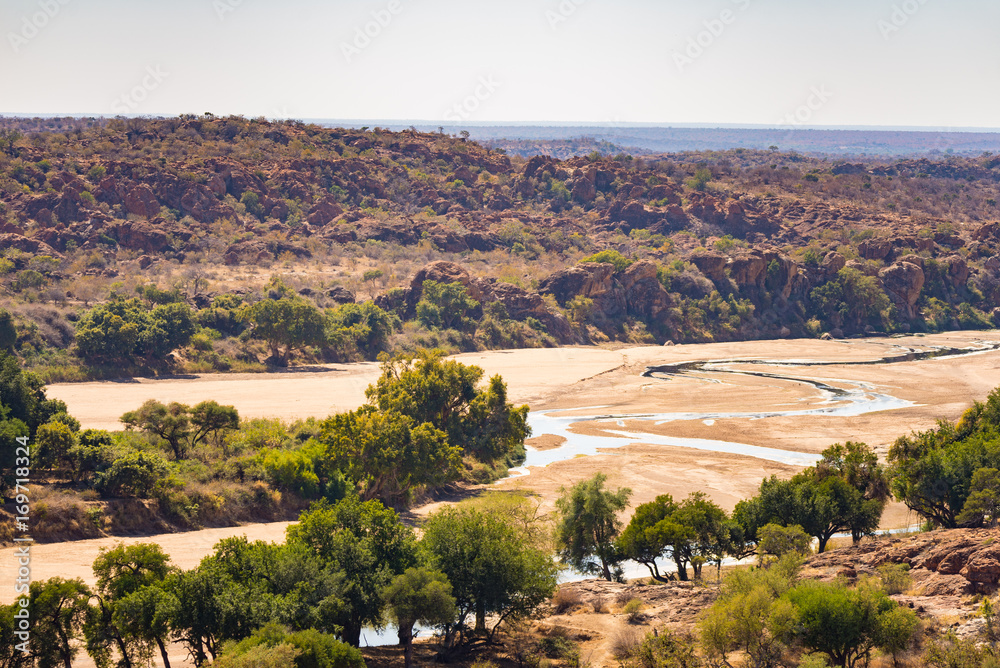 River crossing the desert landscape of Mapungubwe National Park, travel destination in South Africa. Braided Acacia and huge Baobab trees with red sandstone cliffs.