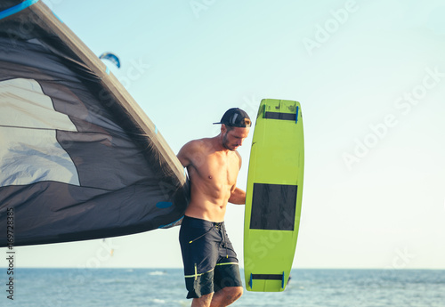 Handsome Caucasian man professional surfer standing on the sandy beach with his kite and board.