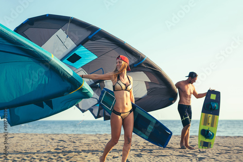 Pretty smiling Caucasian woman and man kitesurfer walking on the beach with their kite
