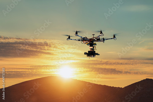The drone with the professional camera takes pictures of the misty mountains at sunset. Uav drone copter flying with digital camera. Hexacopter drone with high resolution digital camera on the sky. © Alexey Yuzhakov