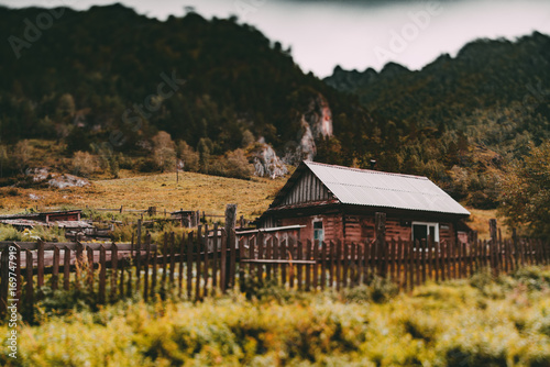 True tilt-shift view of old wooden rustic home in mountains settings with fence in foreground and forest on flank of hill in defocused background, autumn day, Altai mountains in Kuyus district, Russia