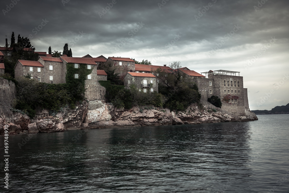 Old ancient buildings on Sveti Stefan island surrounded by sea water on coastline during sunset  with dramatic sky in Europe country Montenegro in fall season