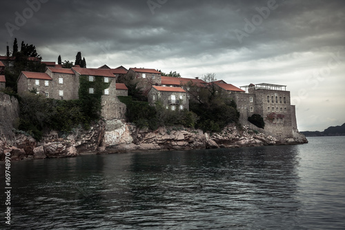 Old ancient buildings on Sveti Stefan island surrounded by sea water on coastline during sunset with dramatic sky in Europe country Montenegro in fall season