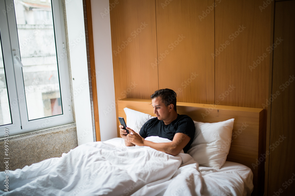 Young man in bed waking up with cellphone at home in the morning Young man in bed waking up with cellphone at home in the morning