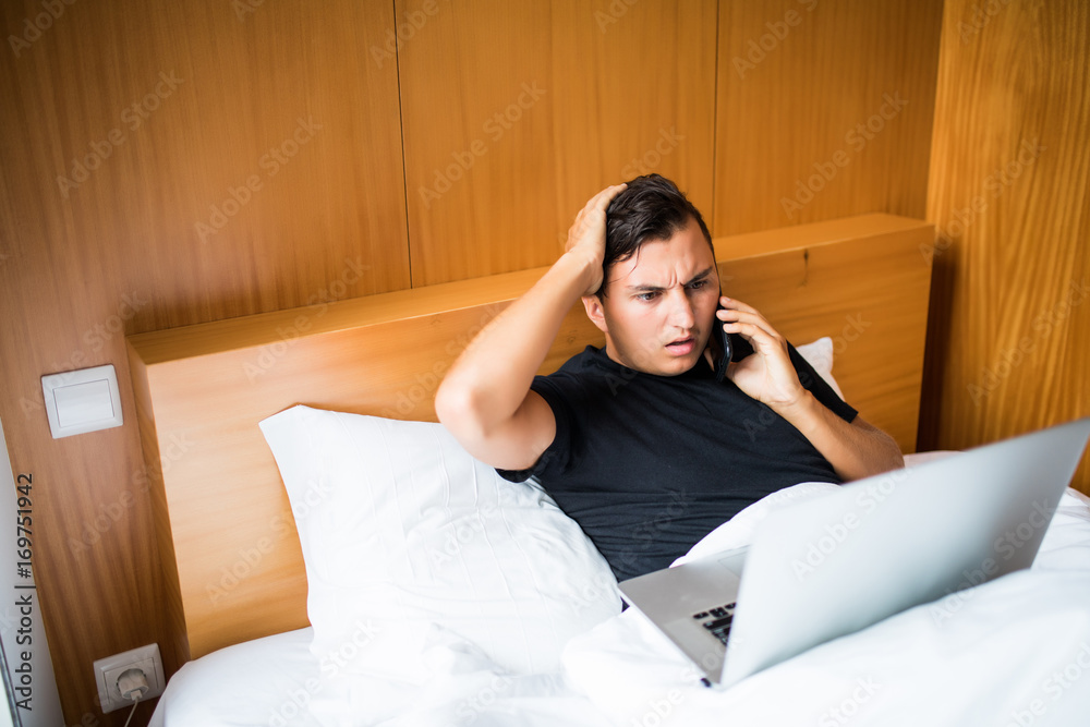 Handsome man lying in morning bed working with laptop and talk phone get surprise news Handsome man lying in morning bed working with laptop and talk phone get surprise news
