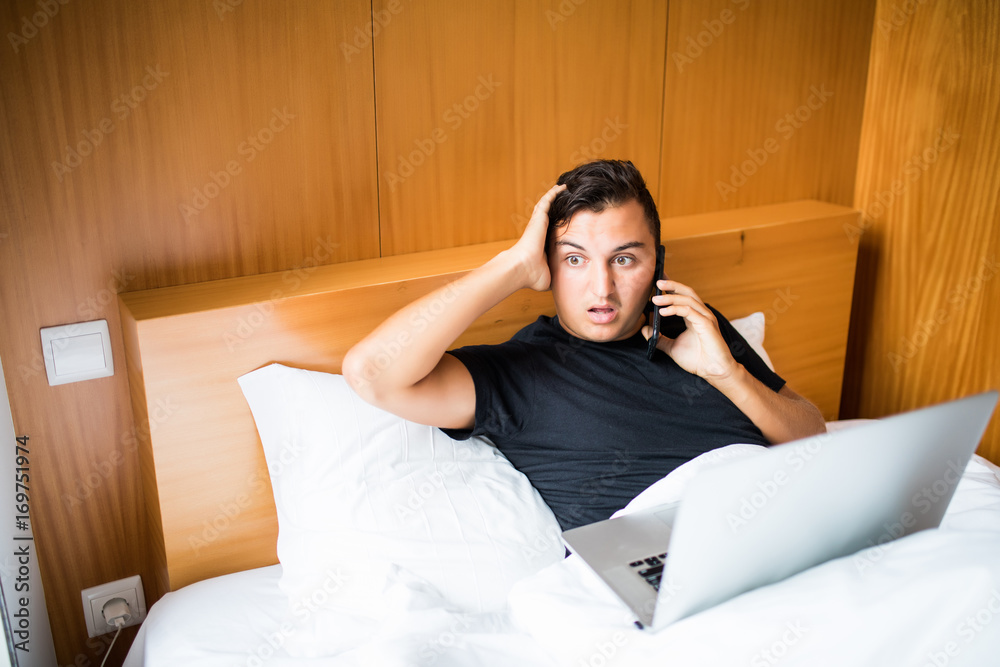 Handsome man lying in morning bed working with laptop and phone shocked reaction Handsome man lying in morning bed working with laptop and phone shocked reaction