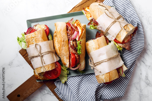 Fresh baguette sandwich bahn-mi styled. Bacon, roasted cheese, tomatoes and lettuce on metallic tray on white marble background.