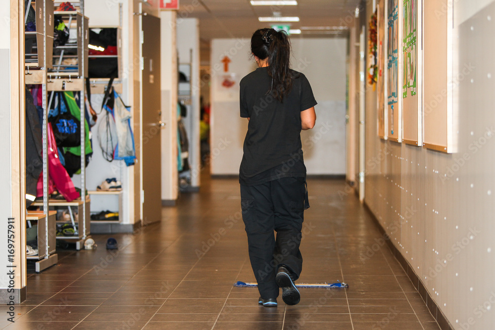 Obraz premium A woman, seen from behind, is sweeping and cleaning the floors in a school corridor.