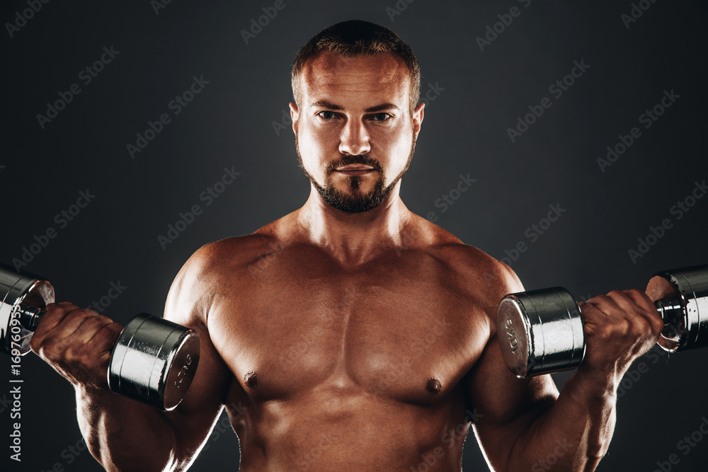 Strong athletic bodybuilder doing exercises with dumbbell,  six pack, perfect abs, shoulders, biceps, and triceps  chest, personal fitness trainer flexing his muscles