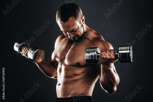 Strong athletic bodybuilder doing exercises with dumbbell, six pack, perfect abs, shoulders, biceps, and triceps chest, personal fitness trainer flexing his muscles