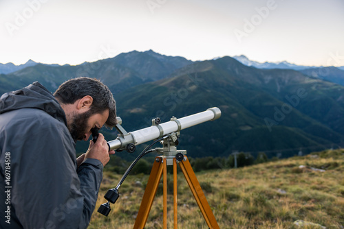Man looking through telescope mountain peaks in summer evening at sunset on mountain outdoor.