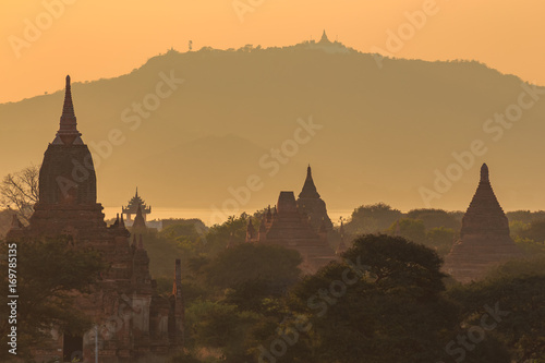 View from afar of the ancient pagodas  stupas  visible among rugged fields and trees of other pagodas and mountains on the horizon during sunset or sunrise  in Bagan  Myanmar  Burma 