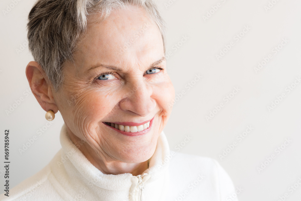 Close up of beautiful older woman with short grey hair and blue eyes in cream jacket laughing against neutral background (selective focus) Close up of beautiful older woman with short grey hair and blue eyes in cream jacket laughing against neutral background (selective focus)