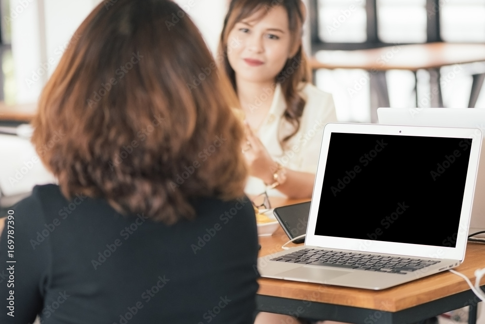 Blank screen laptop place on wooden table. Asian woman working with her friend in coffee shop in selective focus. Blank screen laptop place on wooden table. Asian woman working with her friend in coffee shop in selective focus.