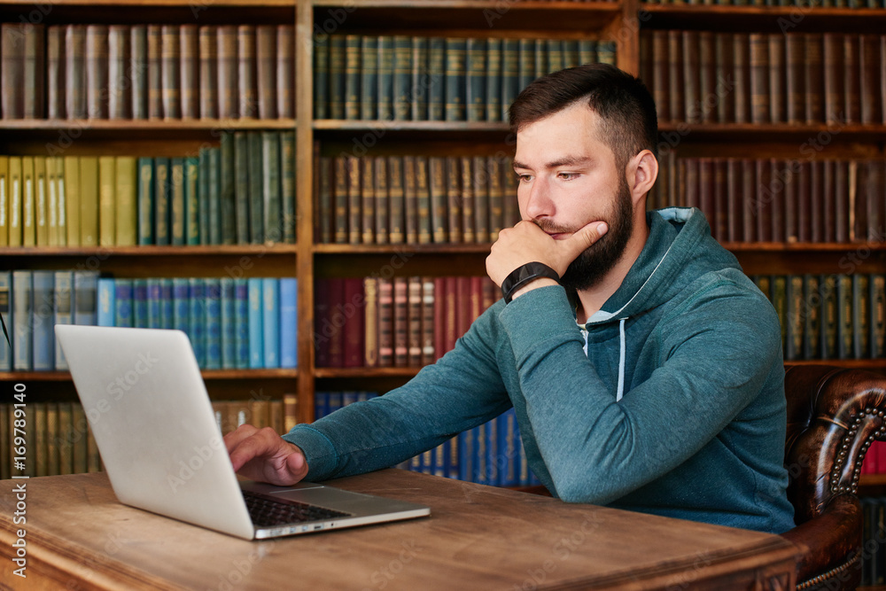The guy sitting at the table with laptop on background of books The guy sitting at the table with laptop on background of books