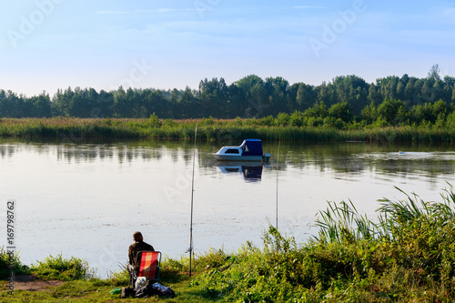 Fishing in the morning - boat on the water, man sitting on the fisherman's chair on the shore of the river