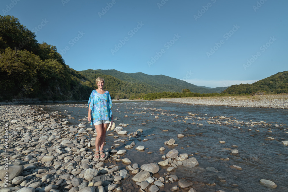 Blond woman is standing in the valley of the mountain river on a background of mountains