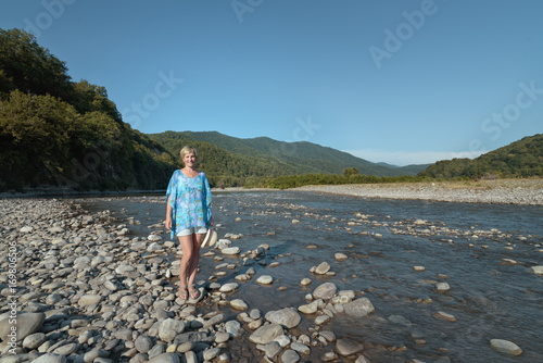 Blond woman is standing in the valley of the mountain river on a background of mountains © alexx_60