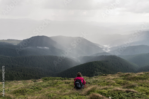 Young female hiker with backpack sits in grass on top of hill. Traveller on trip to mountains taking rest at mountain peak and looking at heavy thunder rain falling on nearby forests