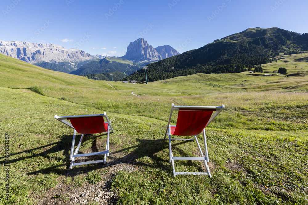 Pleasant holiday in the Dolomites, Italy. Relaxation in the mountains - summer mountains landscape. Two red easy chairs on the meadow in the Alps. Pleasant holiday in the Dolomites, Italy. Relaxation in the mountains - summer mountains landscape. Two red easy chairs on the meadow in the Alps.