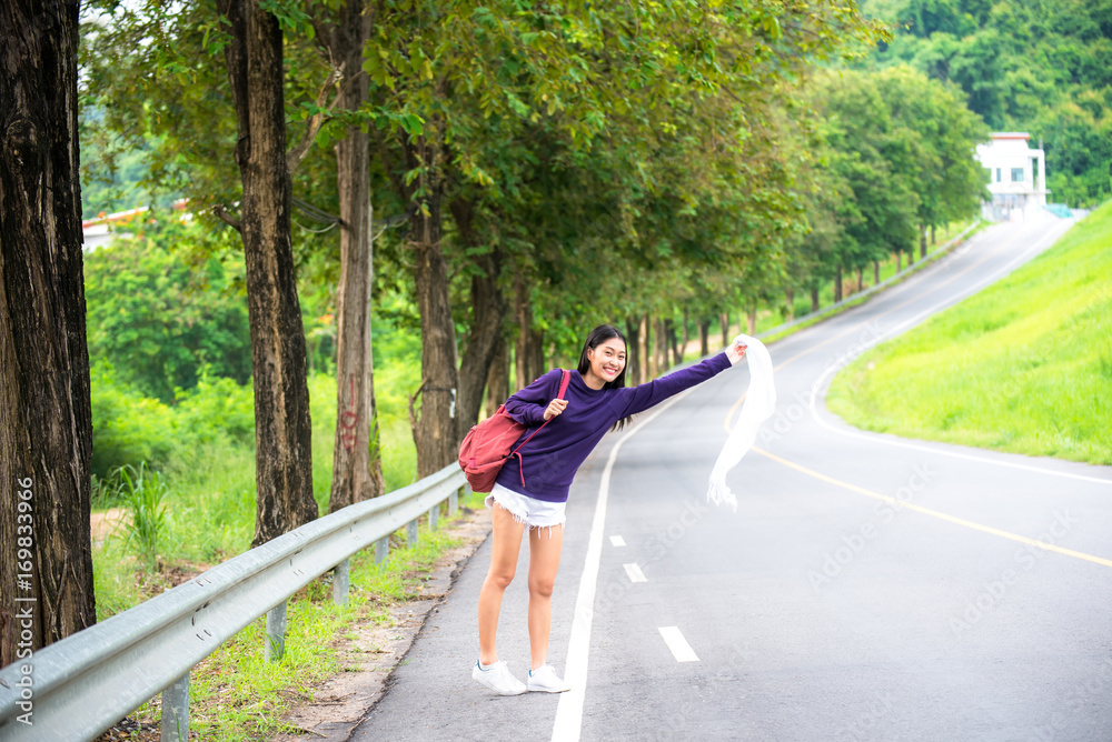 Happy pretty young woman with hand up calling passing car on countryside street,