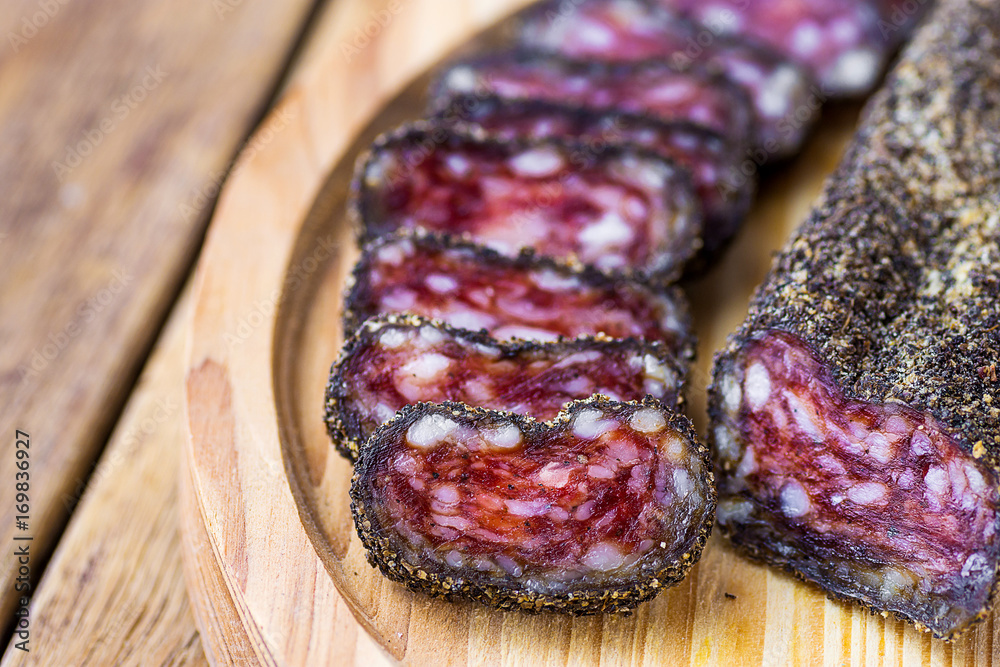 Dry-cured sausage coated with black pepper crust sliced on wood cutting board. Traditional Spanish or mediterranean delicacy. Close up, copy space. Template for poster, menu.