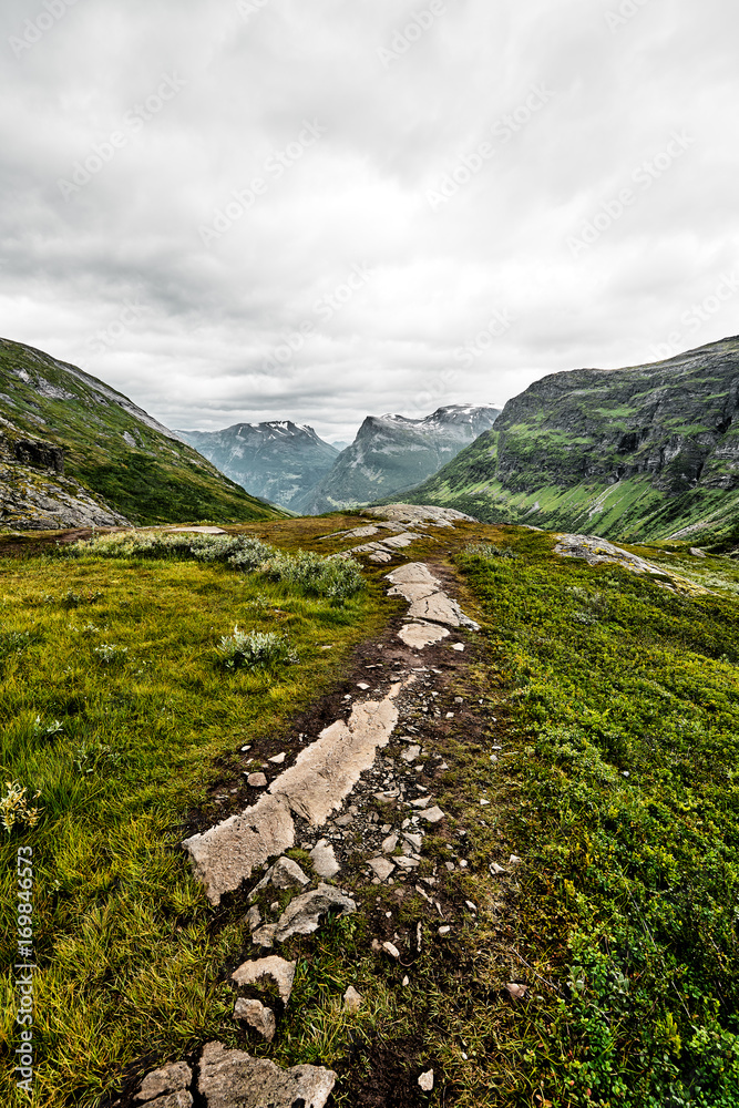 Path over green pasture in the mountains of Western Norway with snow on the summits and a dark cloudy sky