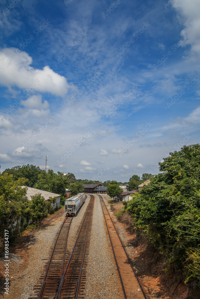Obraz premium Railway lines stretch away and turn before a building in the distance. A rail car sits on the siding. Trees and other buildings line the tracks. Wispy clouds are seen in front of a blue sky.