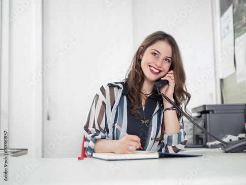 Cheerful young woman is talking on the phone while working at her office photo