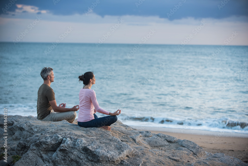 Couple practicing yoga at sunrise on the beach facing the sea