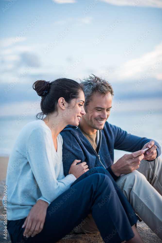Obraz premium A couple sitting at the beach, looking at photos on a phone