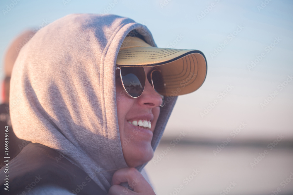 Close portrait of young smiling woman wearing cap, hood and sunglasses. Blurred lake or river on the background, travel, expedition concept.