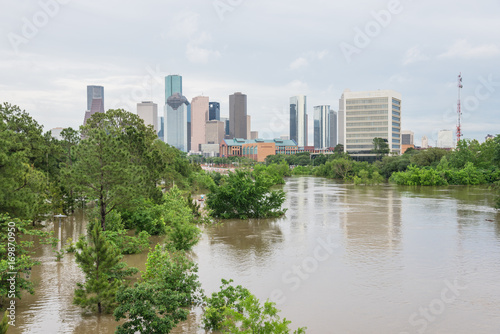 High and fast water rising in Bayou River along Allen Parkway and Memorial Drive with downtown Houston in background under storm cloud sky. Heavy rains from tropical storm caused many flooded areas