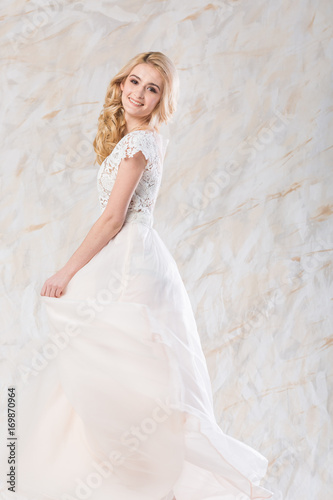 fashionable wedding dress, beautiful blonde model, bride hairstyle and makeup concept - happy young woman in long luxury white gown indoors on light background, stunning female posing with a smile © melnikofd