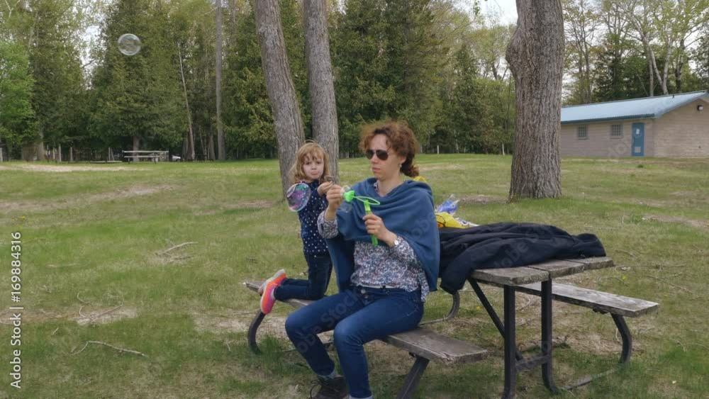 A mother seated on a park bench entertains her daughter by making soap bubbles. Long shot. A mother seated on a park bench entertains her daughter by making soap bubbles. Long shot.
