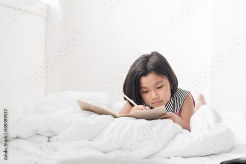Cute Asian girl writing the book while lying on the bed,Learning and education concept
