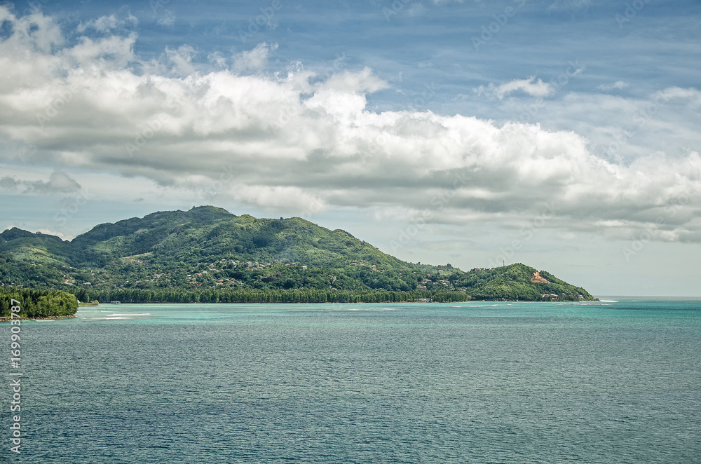 An island with green hills, blue water around and big clouds above, Seychelles