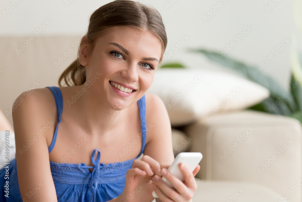 Portrait of beautiful young woman lying on sofa using cellphone looking at camera. Attractive millennial lady with mobile phone in hands calling friends, chatting online, writing message, enjoying app Portrait of beautiful young woman lying on sofa using cellphone looking at camera. Attractive millennial lady with mobile phone in hands calling friends, chatting online, writing message, enjoying app