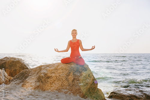 A woman in a red suit practicing yoga on stone at sunrise near the sea