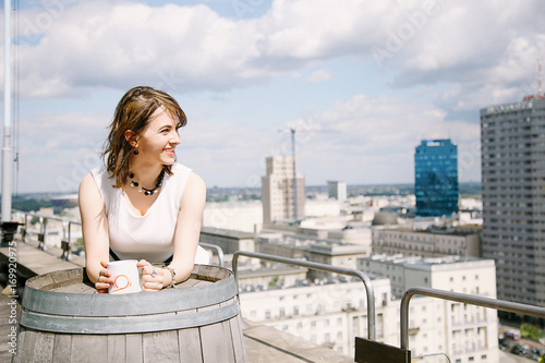 Fototapeta Naklejka Na Ścianę i Meble -  The girl reads a book overlooking the Palace of Culture in Warsaw, a business woman. The lady sits at the table, drinks coffee and relaxes from work. Working break. Fine day.