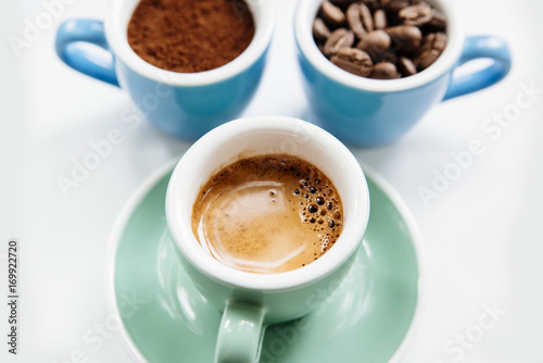 Three cups of coffee: ground coffee (powder), coffee beans and freshly made espresso in colorful ceramic cups (mint green and light blue) on the white background